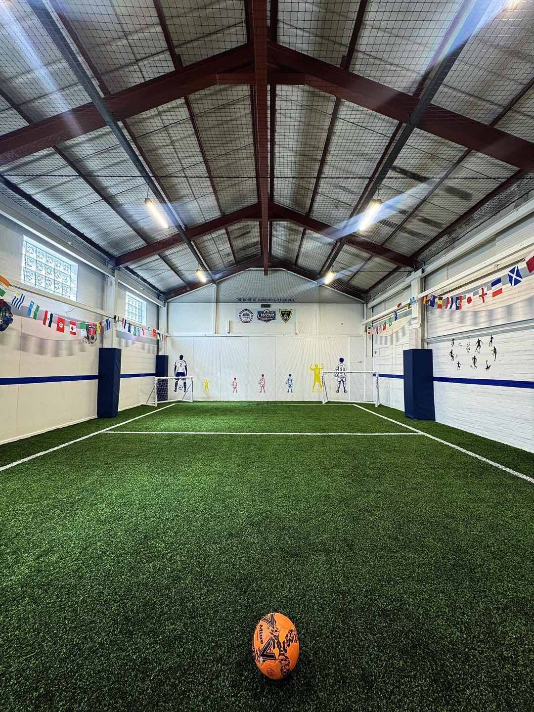 Inside the WDR Football Centre — the astro pitch, goals, and the gable wall lettered “The home of Harborough football”