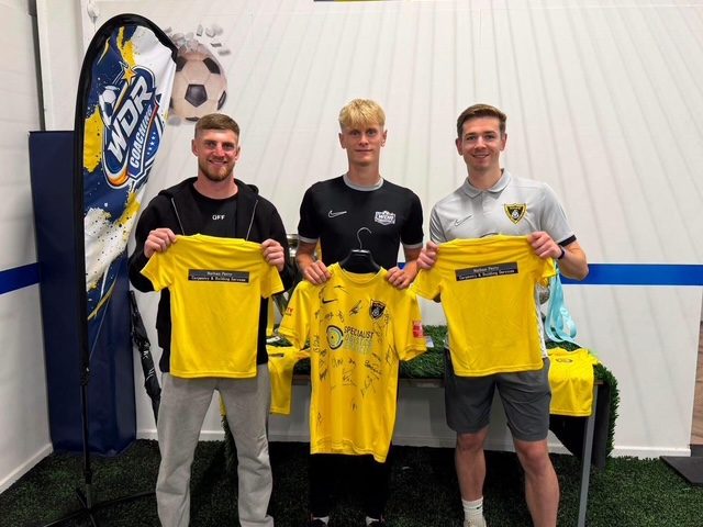 Three Harborough Town players holding up signed yellow club shirts in front of the WDR feather flags