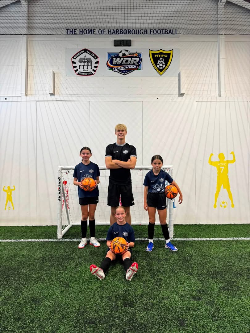 Coach Will with three young players in WDR training tops, each holding a ball, in front of a goal beneath the wall lettered “The home of Harborough football”