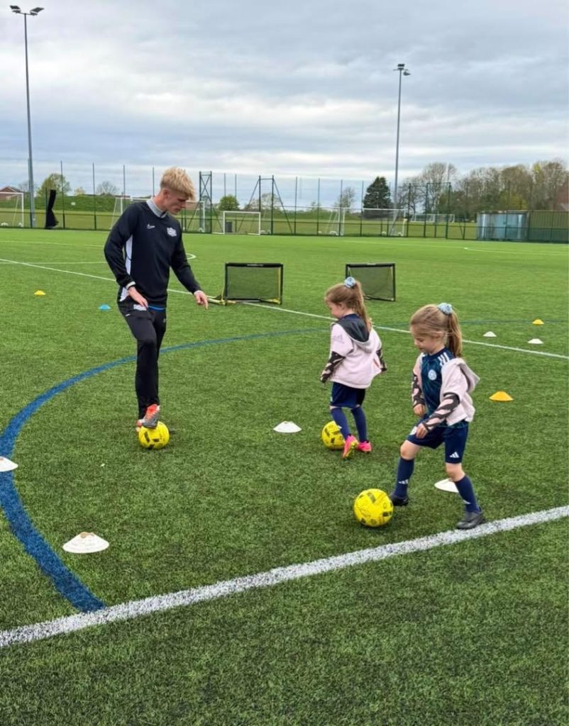 Coach Will running an outdoor session on a 3G pitch, two young players dribbling through cones beside him