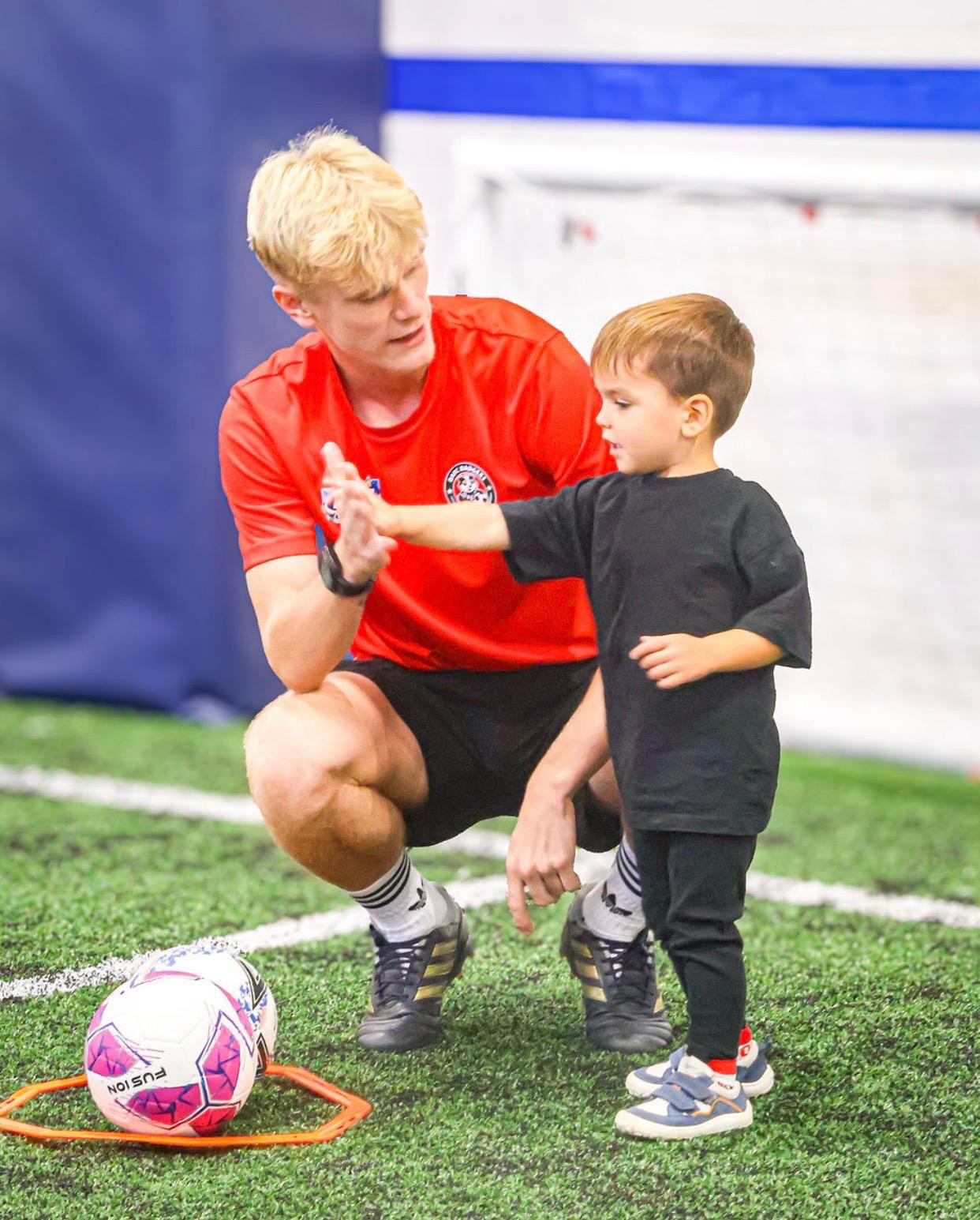Coach Will crouching to high-five a toddler beside a ball at a First Kicks session on the indoor astro pitch