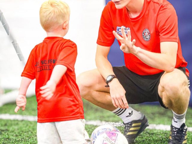 Coach Will crouching to talk to a young player in a red JS Scaffolding First Kicks top on the indoor astro
