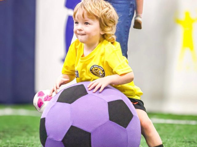 A smiling toddler in a yellow HTFC Bumblebees First Kicks shirt sitting against a giant football