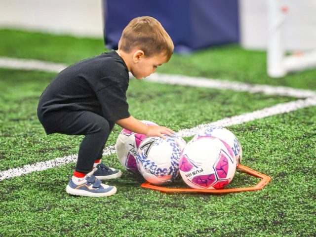 A toddler crouching to pick up footballs from a marker ring on the indoor astro