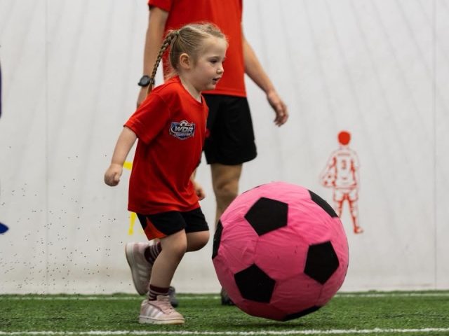 A young girl in a red WDR Coaching top running onto an oversized pink football, with a coach behind her