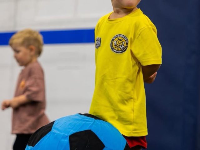 A toddler in a yellow HTFC Bumblebees First Kicks shirt standing over an oversized blue football