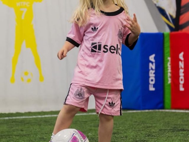A young girl in a pink football kit dribbling a ball across the indoor astro