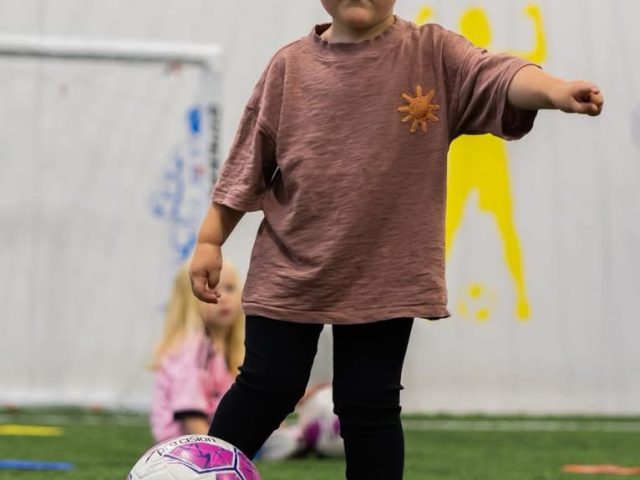 A toddler stepping up to a football on the astro pitch, arm out for balance