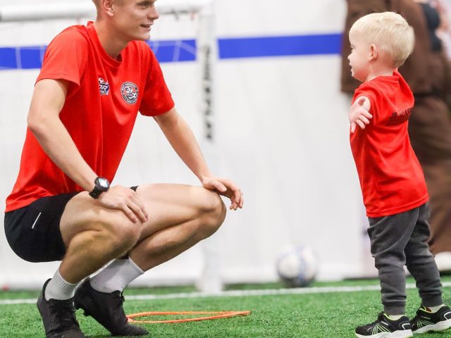 A coach crouched down on the astro talking to a toddler in a red First Kicks shirt