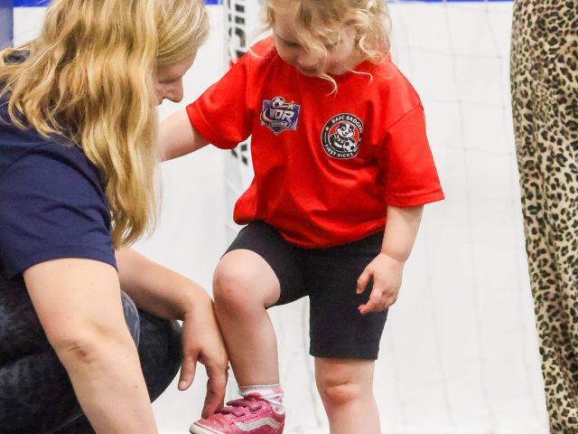 A coach kneeling on the astro to guide a young player's foot onto the ball
