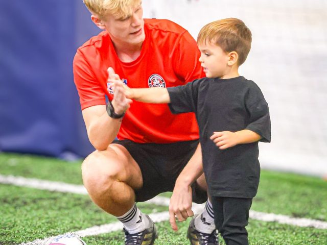 A coach crouching to high-five a young player in black, a ball in a marker ring beside them