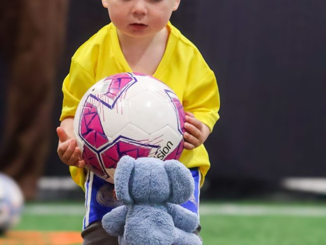 A toddler in a yellow First Kicks shirt lifting a pink ball, a toy elephant on the ball below