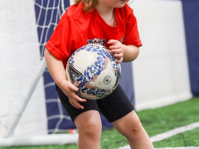 A young girl in a red BAFC Badgers First Kicks shirt carrying a football away from the goal
