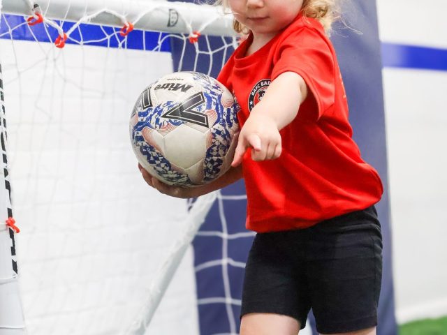 A young girl in a red BAFC Badgers shirt holding a Mitre ball, arm out for balance