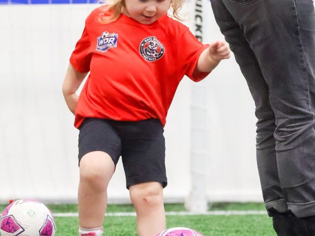 A young girl in a red BAFC Badgers shirt striking a pink football off a marker