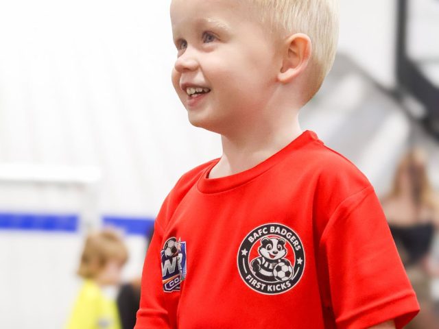 A young boy in a red BAFC Badgers First Kicks shirt smiling across the arena