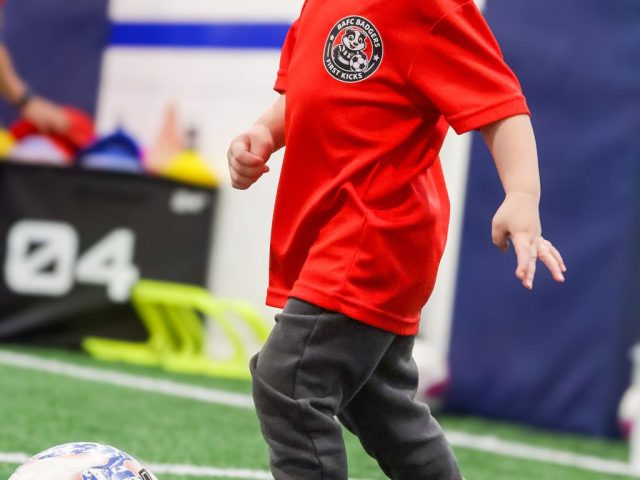 A boy in a red BAFC Badgers First Kicks shirt walking the ball forward