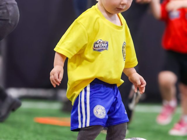 A toddler in a yellow HTFC Bumblebees shirt stepping up to a Mitre ball inside an orange ring
