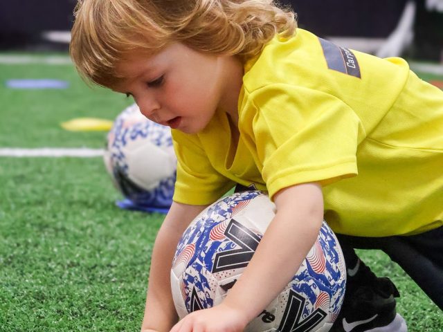A boy in a yellow First Kicks shirt pressing a red marker flat beside his football