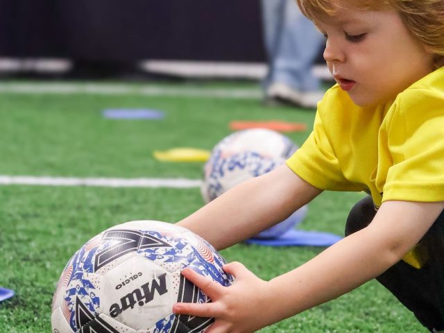 A boy in a yellow First Kicks shirt settling a Mitre ball onto a red marker