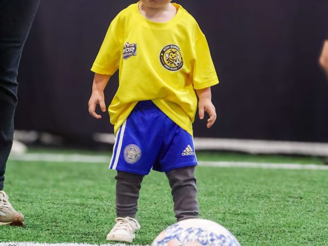A toddler in a yellow HTFC Bumblebees First Kicks shirt standing over a football on the indoor astro