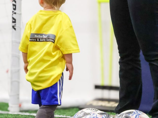 A toddler in a yellow First Kicks shirt stood by the goal, footballs waiting in a marker ring