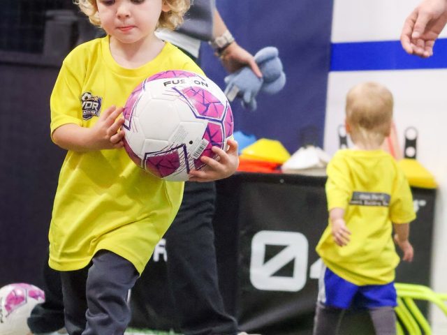 A boy in a yellow First Kicks shirt carrying a pink football under his arm