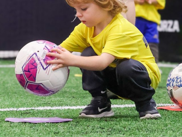 A boy in a yellow First Kicks shirt crouching to pick up a pink football