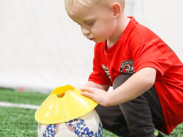 A boy in a red BAFC Badgers shirt balancing a yellow marker cone on top of a football