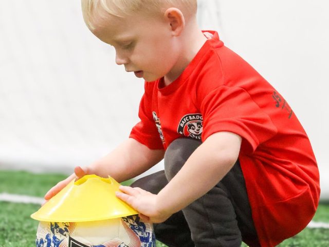 A boy in a red BAFC Badgers shirt setting a yellow cone down on top of a Mitre ball