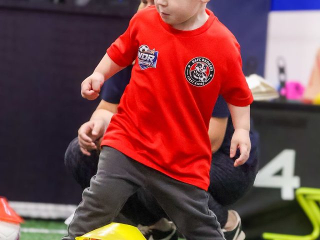 A boy in a red BAFC Badgers shirt dribbling a pink ball over a yellow marker cone