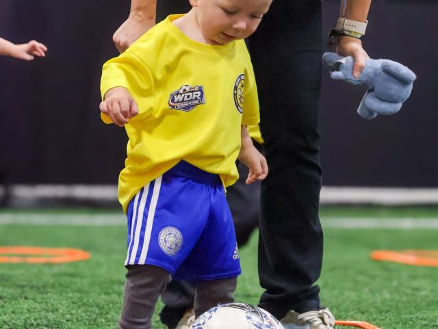A toddler steadied by a coach as he takes a first touch inside a ring of orange markers