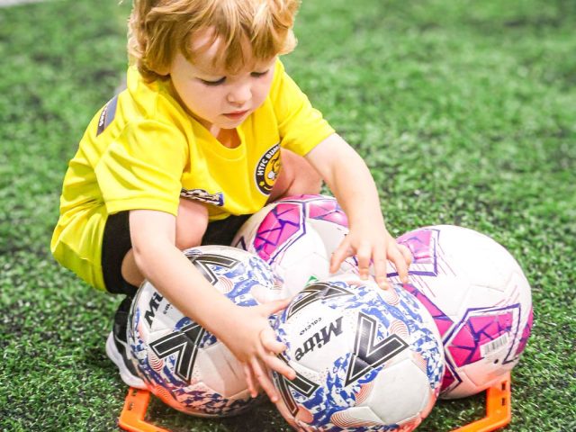A toddler in a yellow First Kicks shirt crouching over a nest of footballs