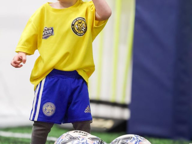 A toddler in a yellow HTFC Bumblebees shirt, hand to his head, two footballs at his feet