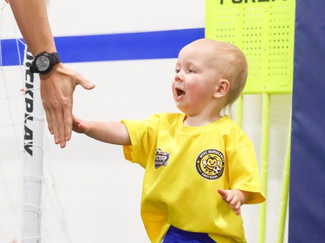 A toddler in a yellow HTFC Bumblebees shirt reaching up to high-five a coach, beside a FORZA free-kick mannequin