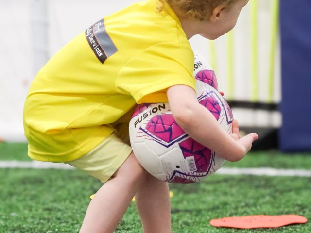 A toddler in a yellow First Kicks shirt crouching to lift a pink football