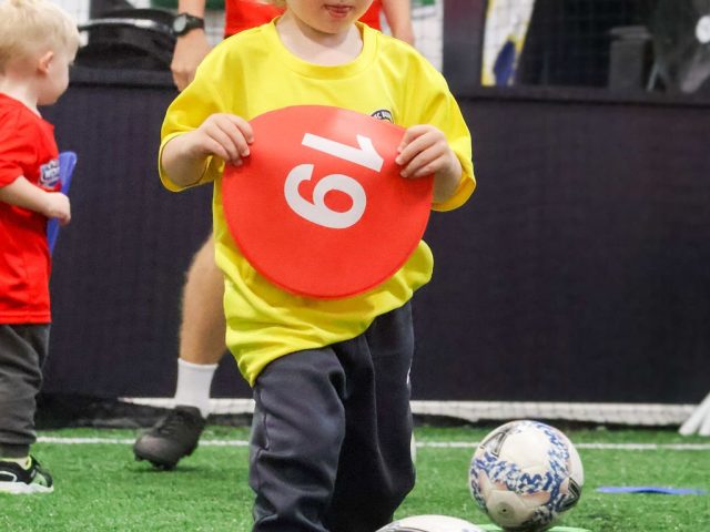 A toddler in a yellow First Kicks shirt holding up a red marker disc numbered 19