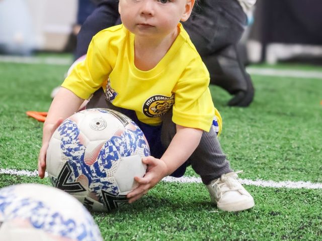 A toddler in a yellow HTFC Bumblebees shirt crouching to pick up a football