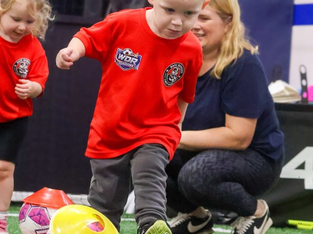 A boy in a red First Kicks shirt running in to kick a ball off a yellow cone, a parent watching behind