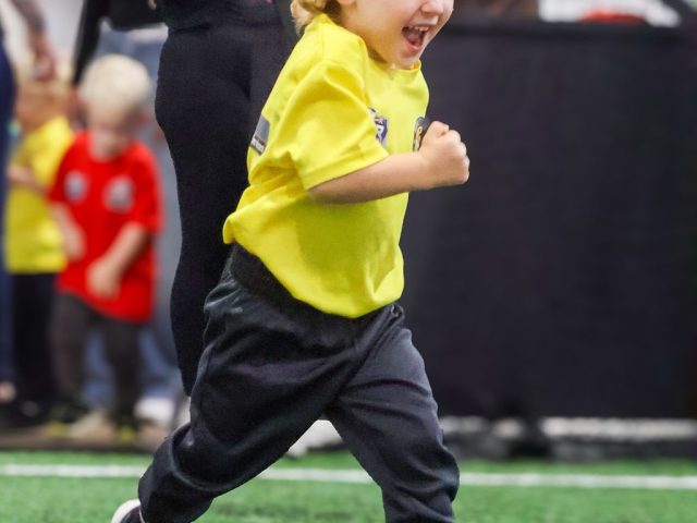 A young boy in a yellow WDR Coaching First Kicks shirt sprinting across the astro, laughing, with parents watching from the side