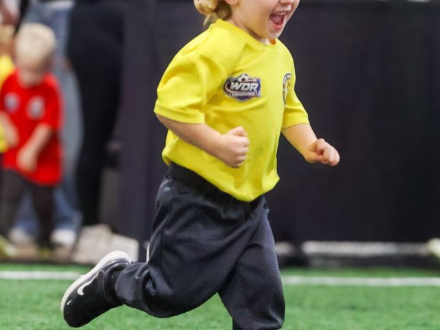 A young boy in a yellow First Kicks shirt running flat out with a huge grin