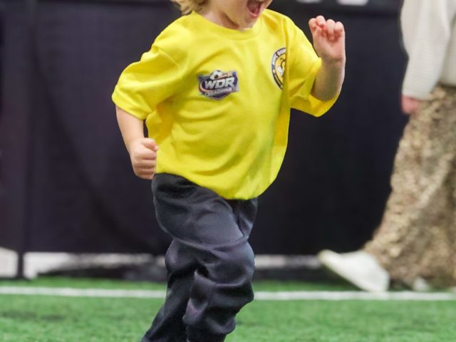 A young boy in a yellow First Kicks shirt running past the side of the pitch, laughing