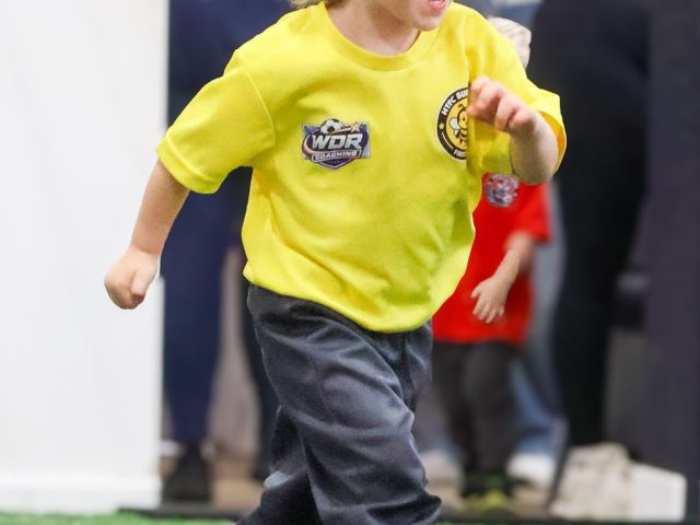 A young boy in a yellow First Kicks shirt running and laughing, mid-stride