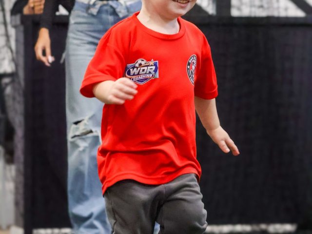 A toddler in a red WDR Coaching First Kicks shirt running across the astro, smiling