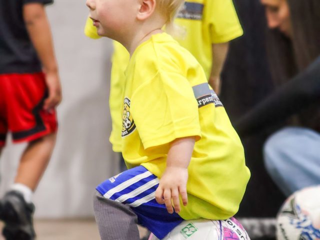 A toddler in a yellow First Kicks shirt perched on a pink football