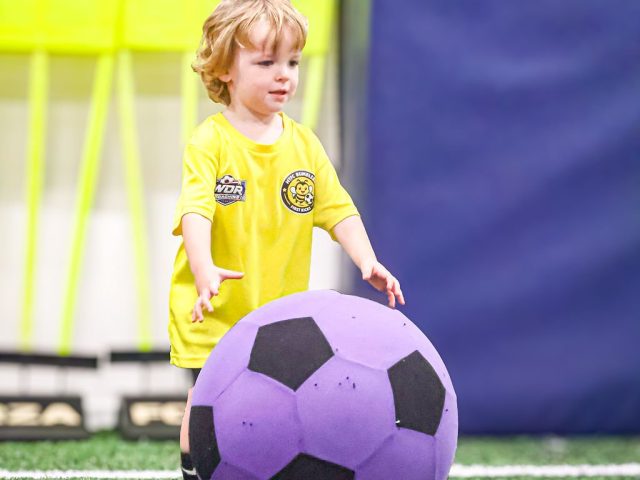 A boy in a yellow HTFC Bumblebees shirt sitting on a football in yellow trainers