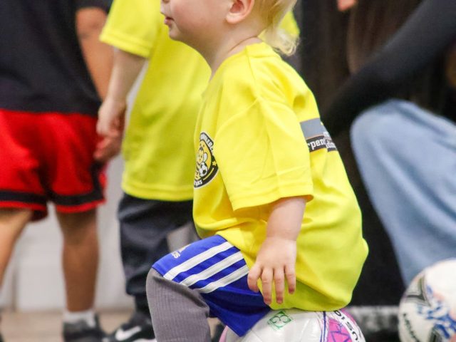 A toddler in a yellow First Kicks shirt resting on a football, watching the session