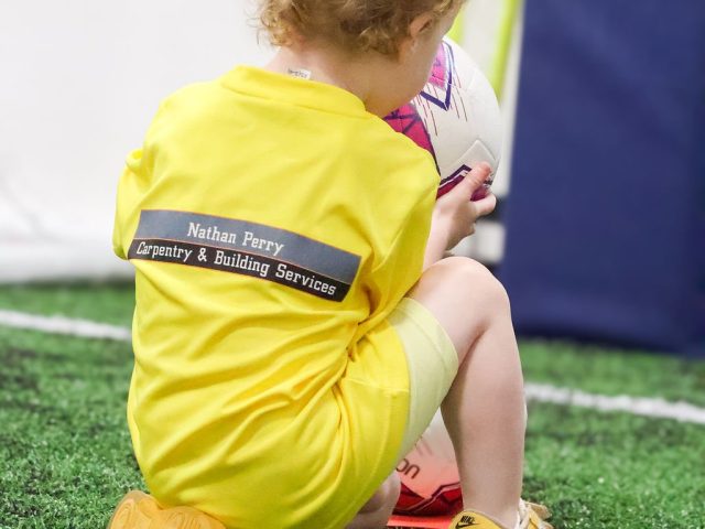 A toddler sat on the astro in a yellow First Kicks shirt backed by Nathan Perry Carpentry and Building Services