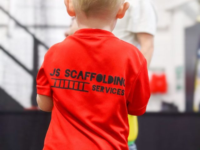 A boy seen from behind in a red First Kicks shirt backed by JS Scaffolding Services