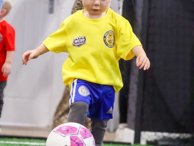 A toddler in a yellow HTFC Bumblebees shirt stepping in to strike a pink football